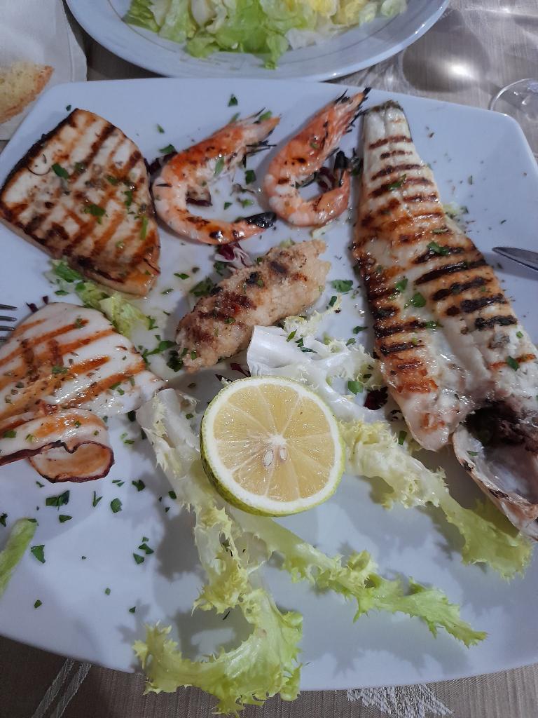 Plate of mixed Italian grilled seafood with swordfish, prawns, squid, fish fillet, and lemon slice.