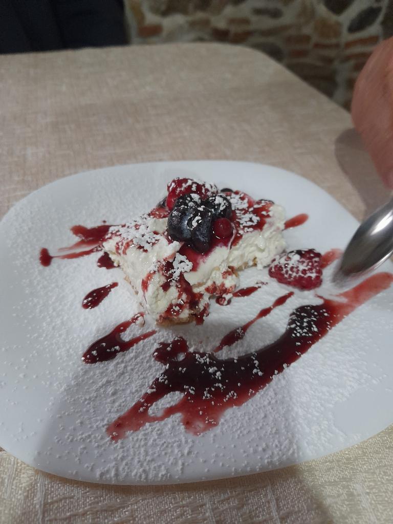 A slice of creamy dessert topped with mixed berries, red berry sauce, and powdered sugar served on a white plate.